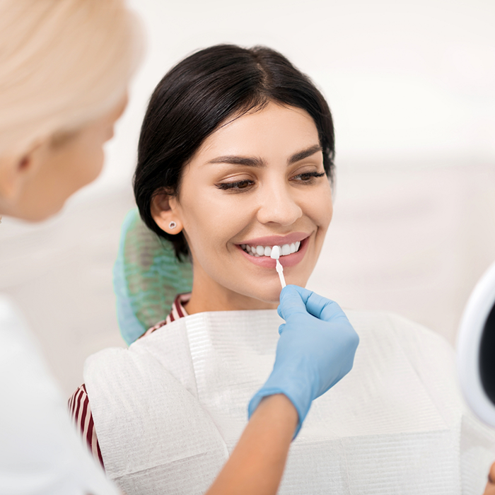 Woman in the dental chair with a dentist holding a veneer in front of her smile